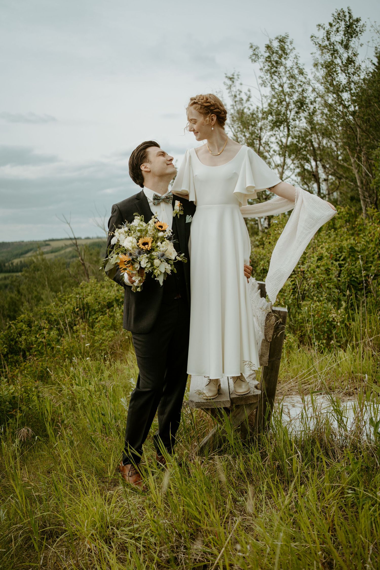 A couple in wedding attire poses on a hilltop meadow with the bride standing on a wooden platform holding a bouquet.