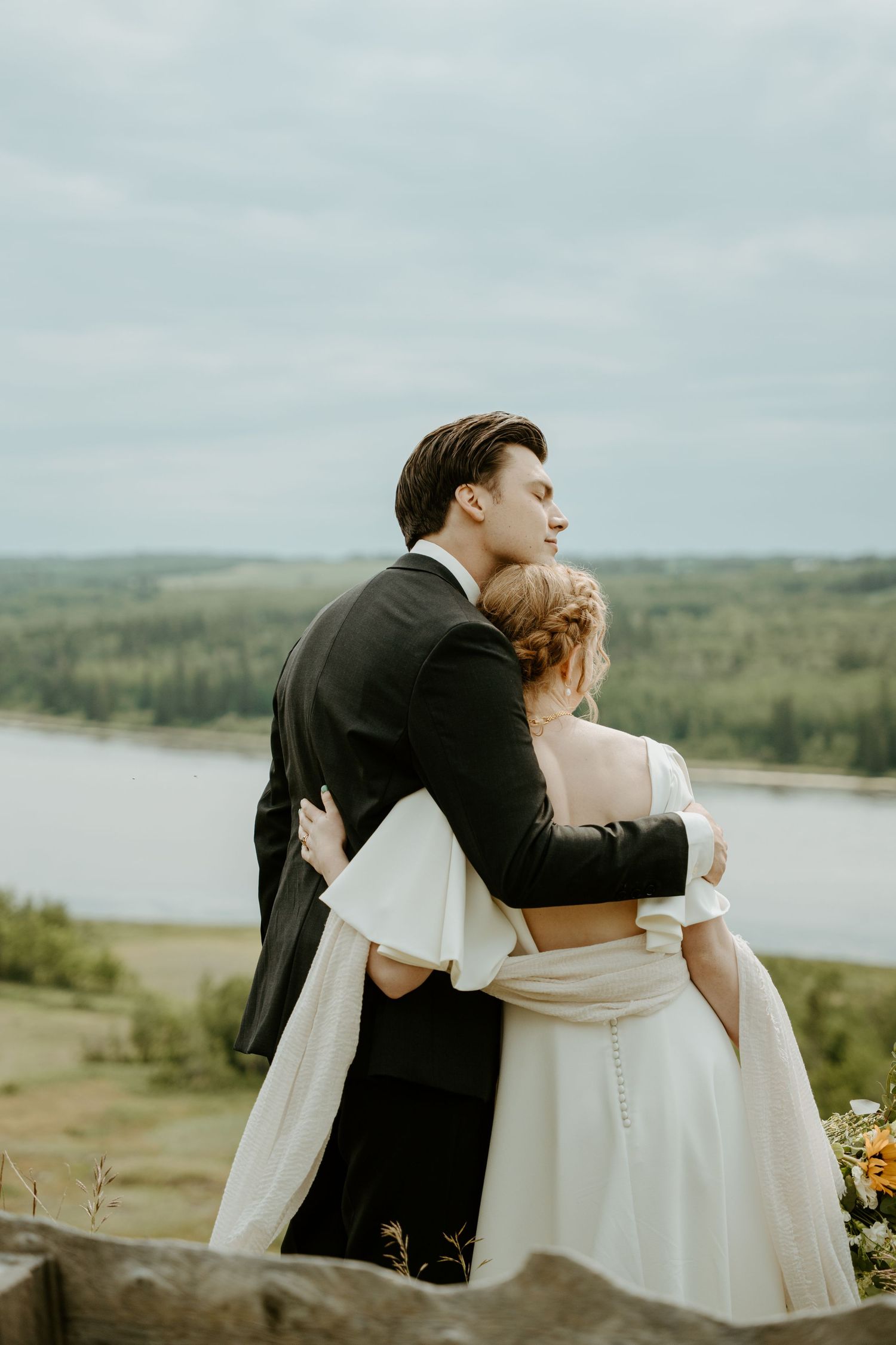 A couple in wedding attire sharing an intimate embrace overlooking a scenic lake and rolling hills on their special day.