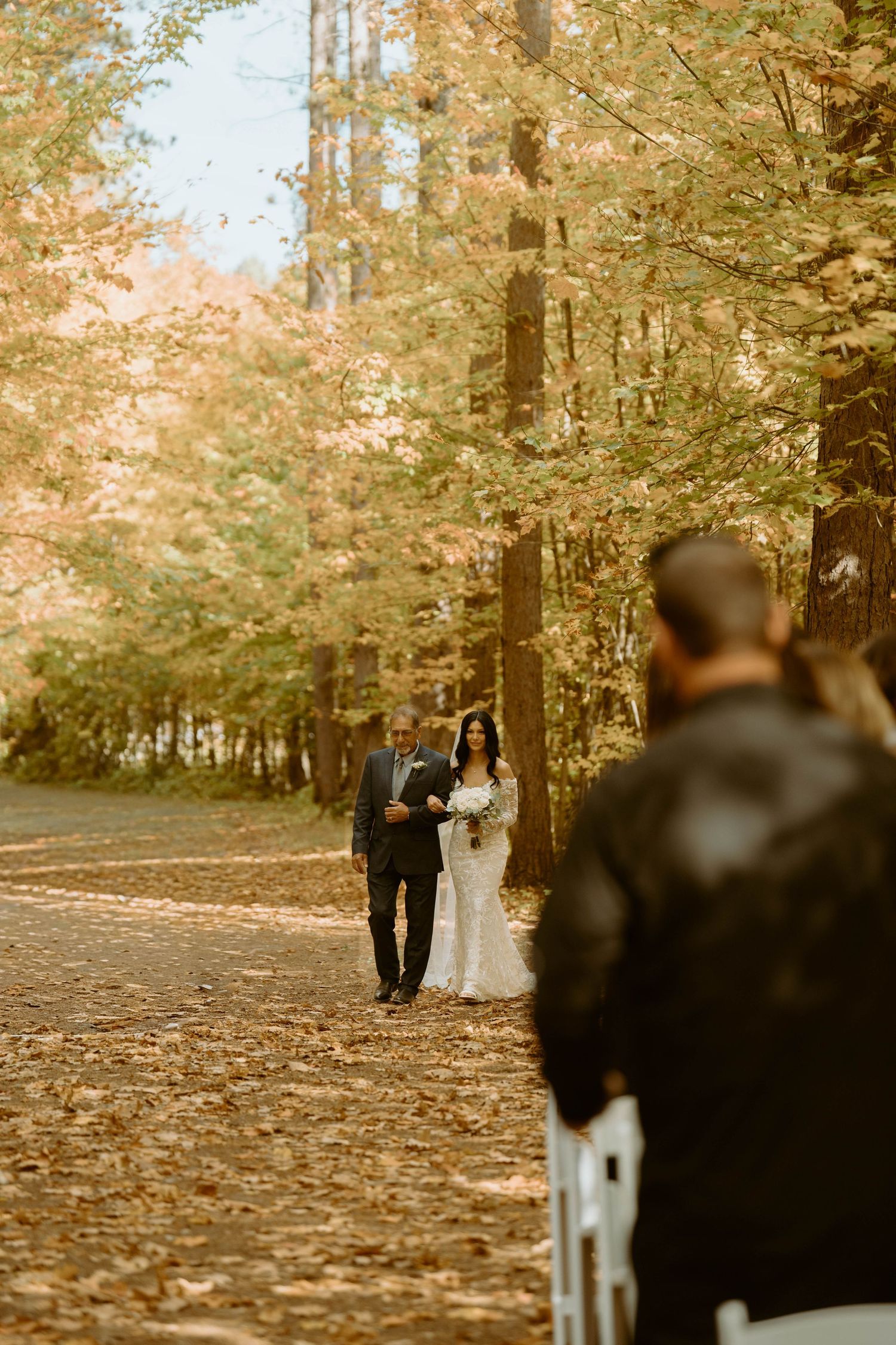 A couple walks through a forest path covered in fallen autumn leaves during a romantic outdoor photo session.