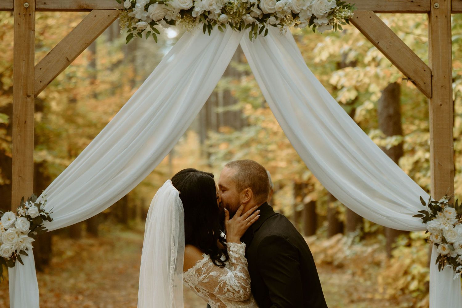 A couple shares a romantic moment under white fabric draping and greenery at an outdoor wedding ceremony.