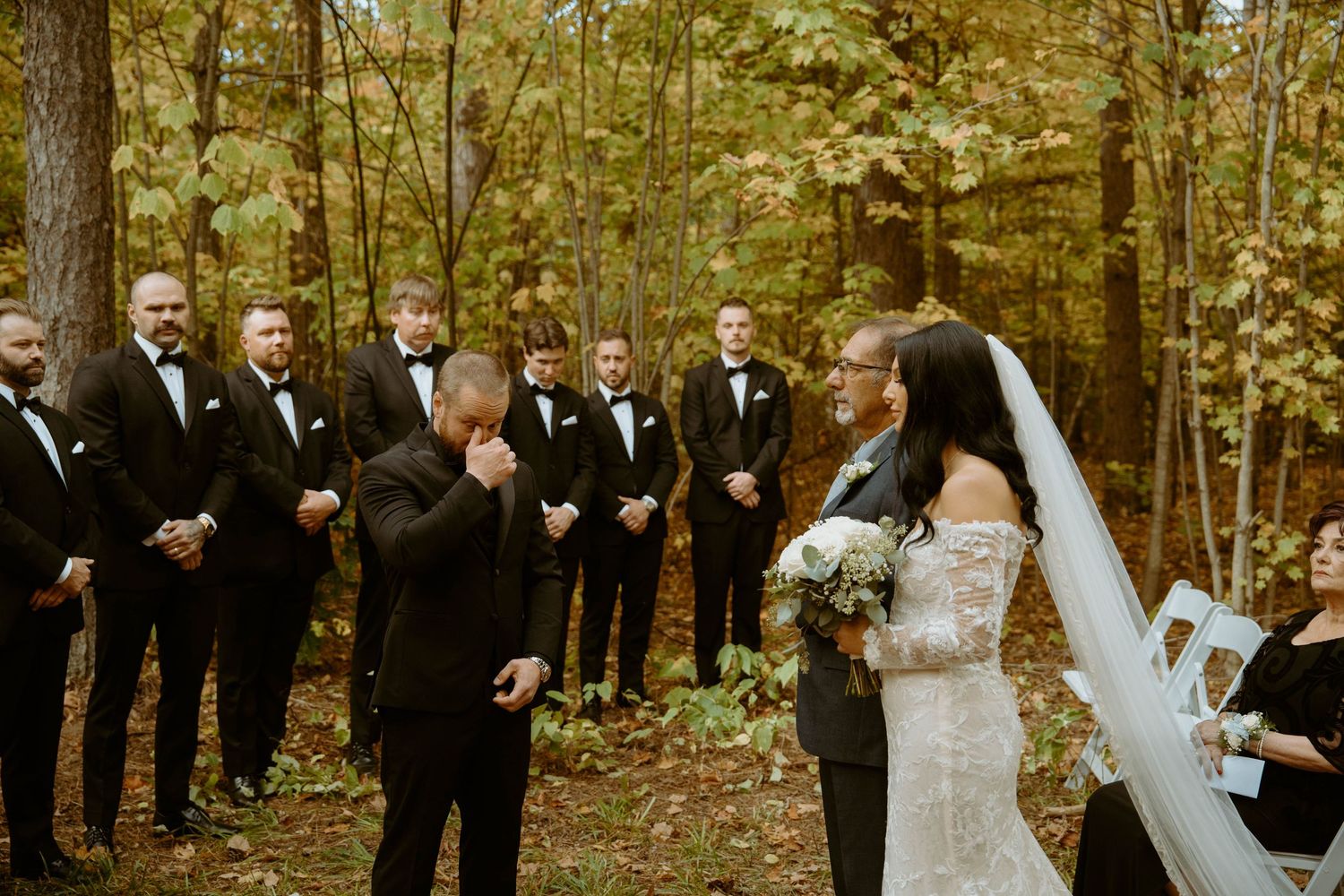 Wedding party in formal attire laughs together during outdoor autumn photo session in wooded setting.
