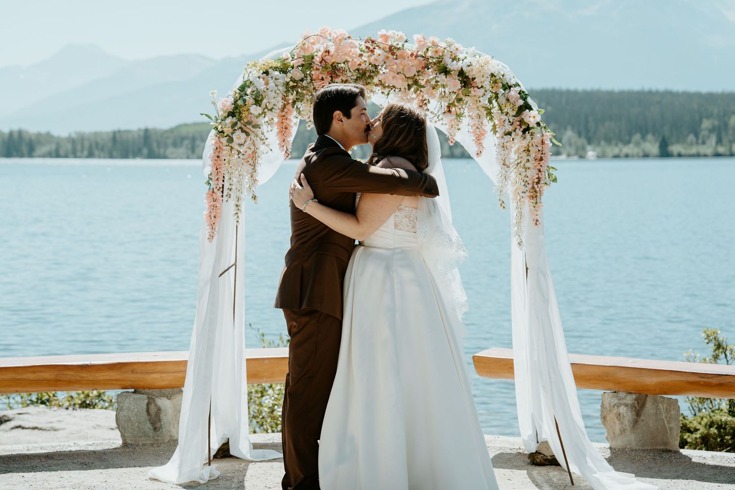 Romantic wedding ceremony setup on a waterfront deck with flowing white fabric and floral decorations overlooking a scenic lake.