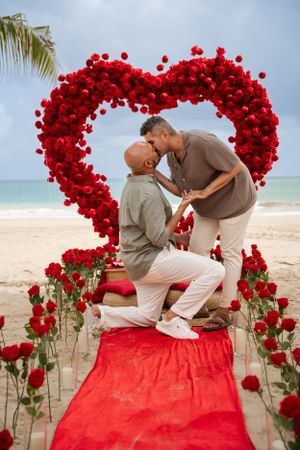 A beach proposal scene with a red heart arch made of roses and a red carpet on the sand.