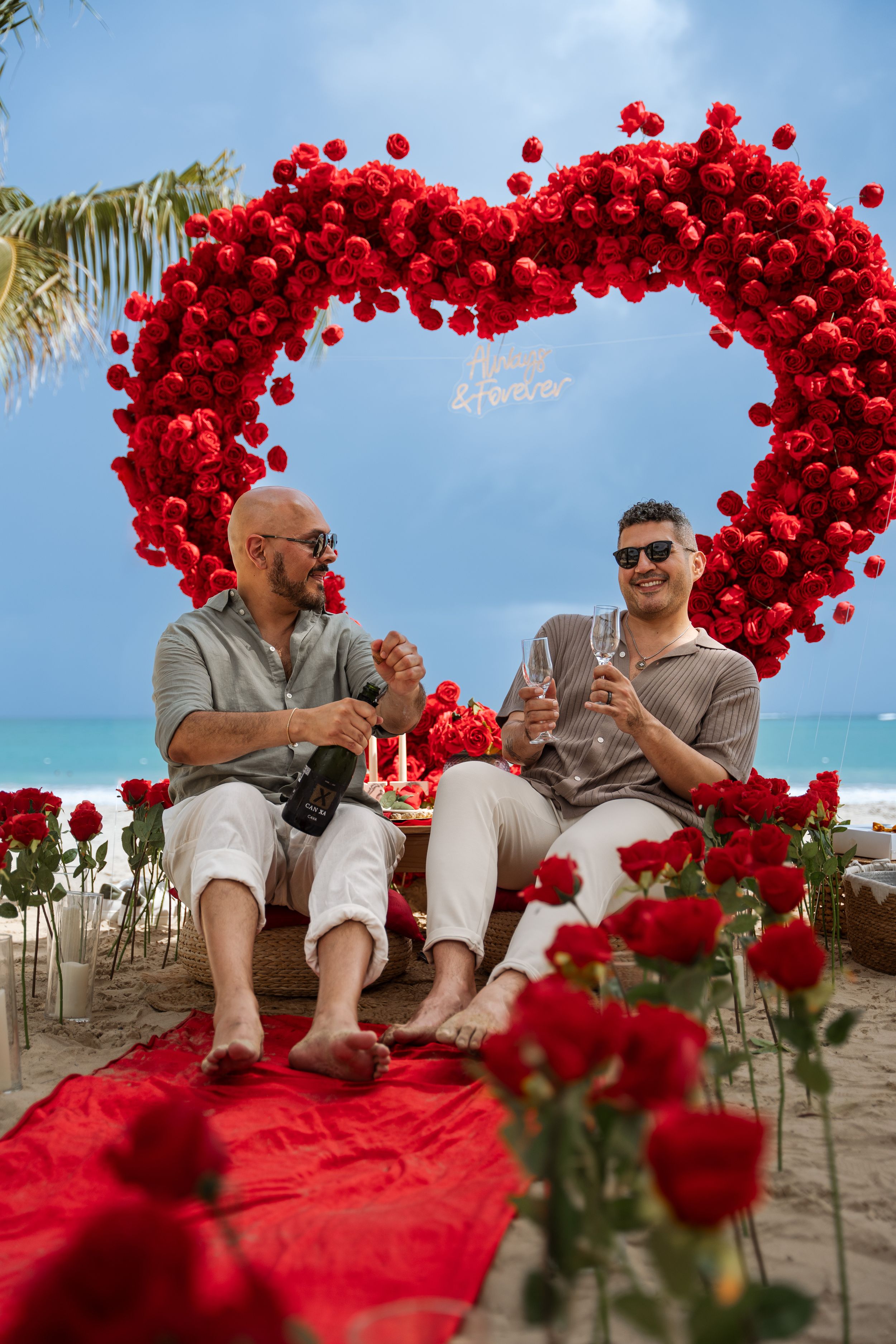 A couple celebrating under a red heart-shaped balloon arch on a tropical beach with roses.