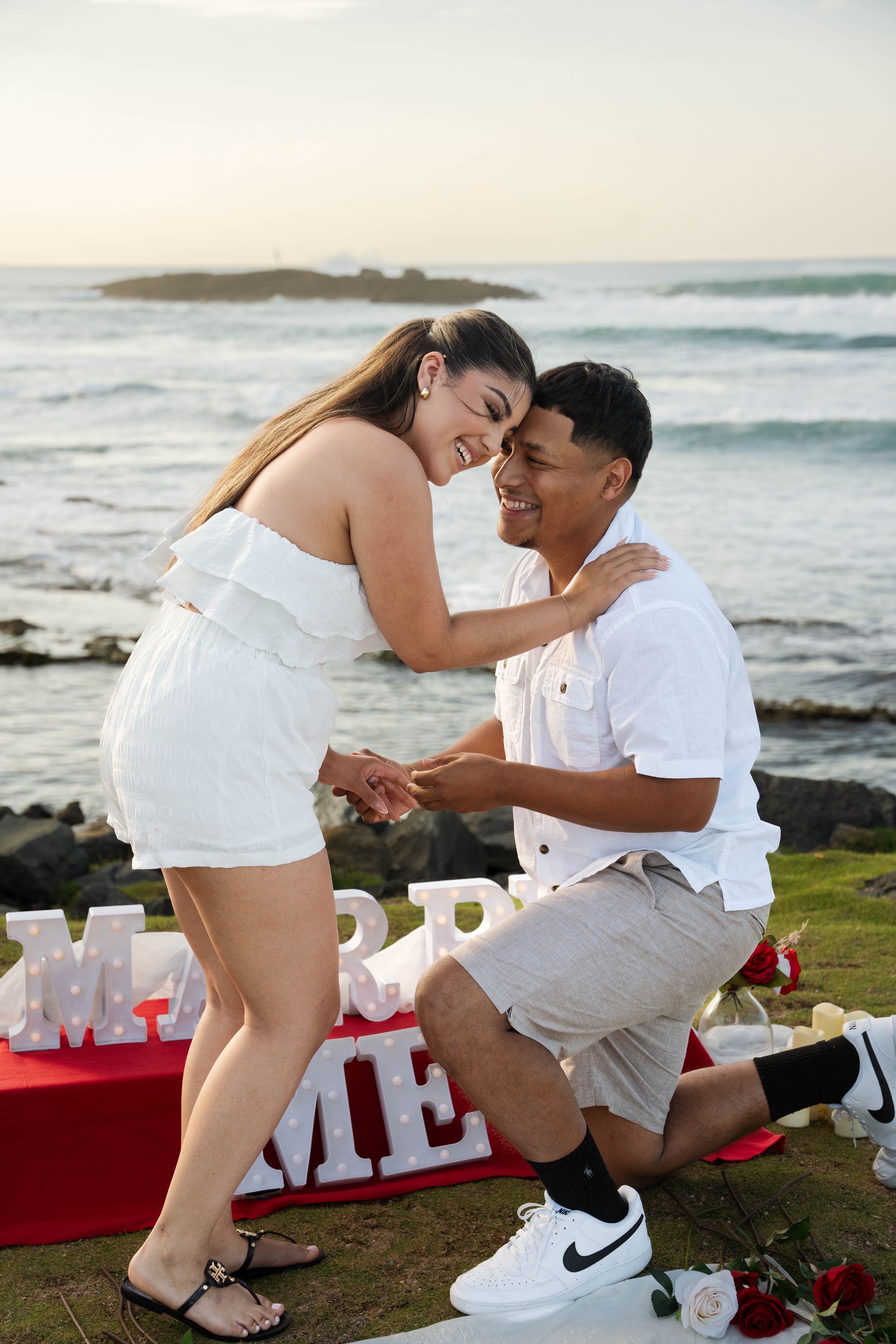 A joyful proposal moment on a scenic coastal cliff with decorative letters and ocean waves in background.