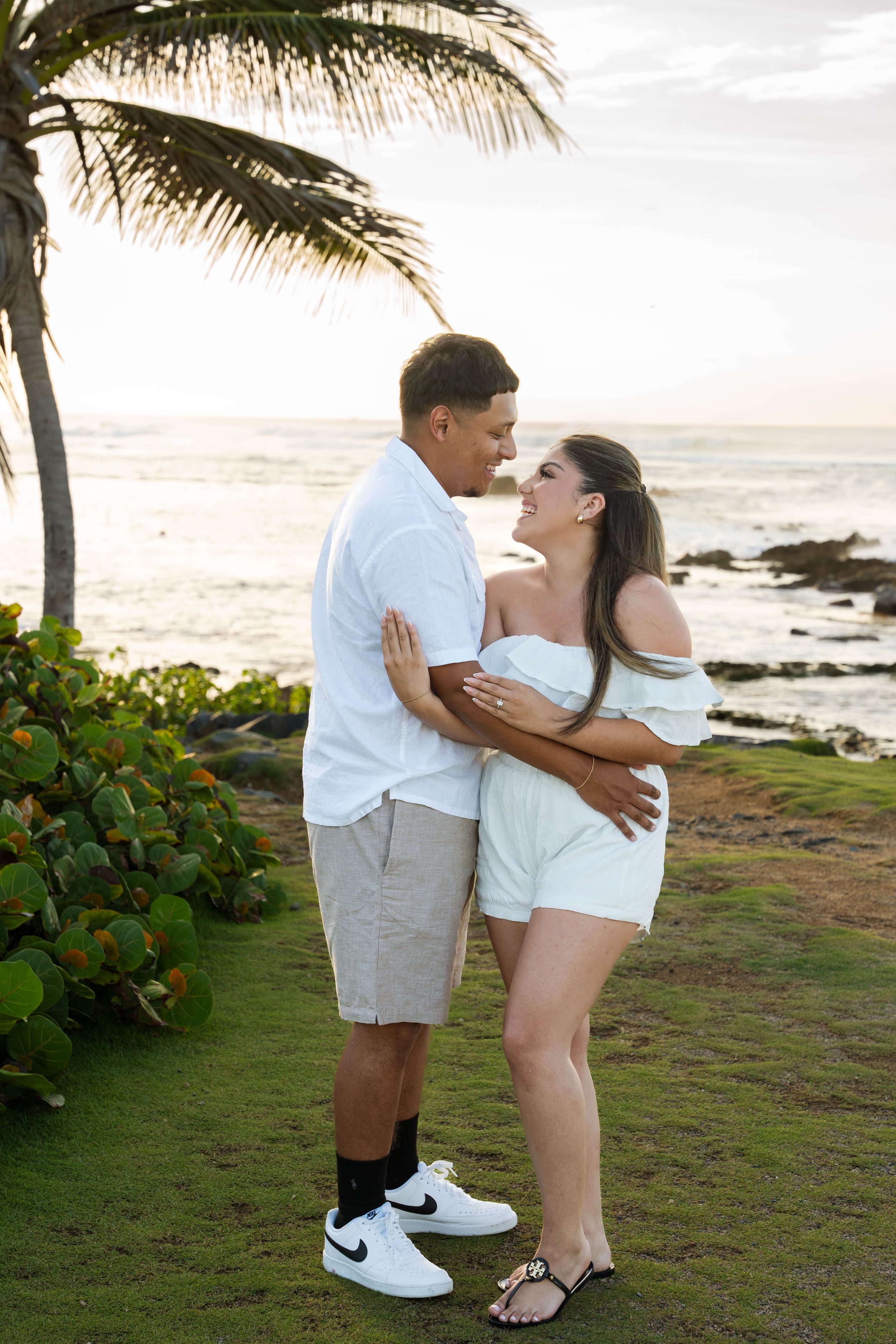 Romantic couple embracing on a beach at sunset with palm trees and ocean in the background.