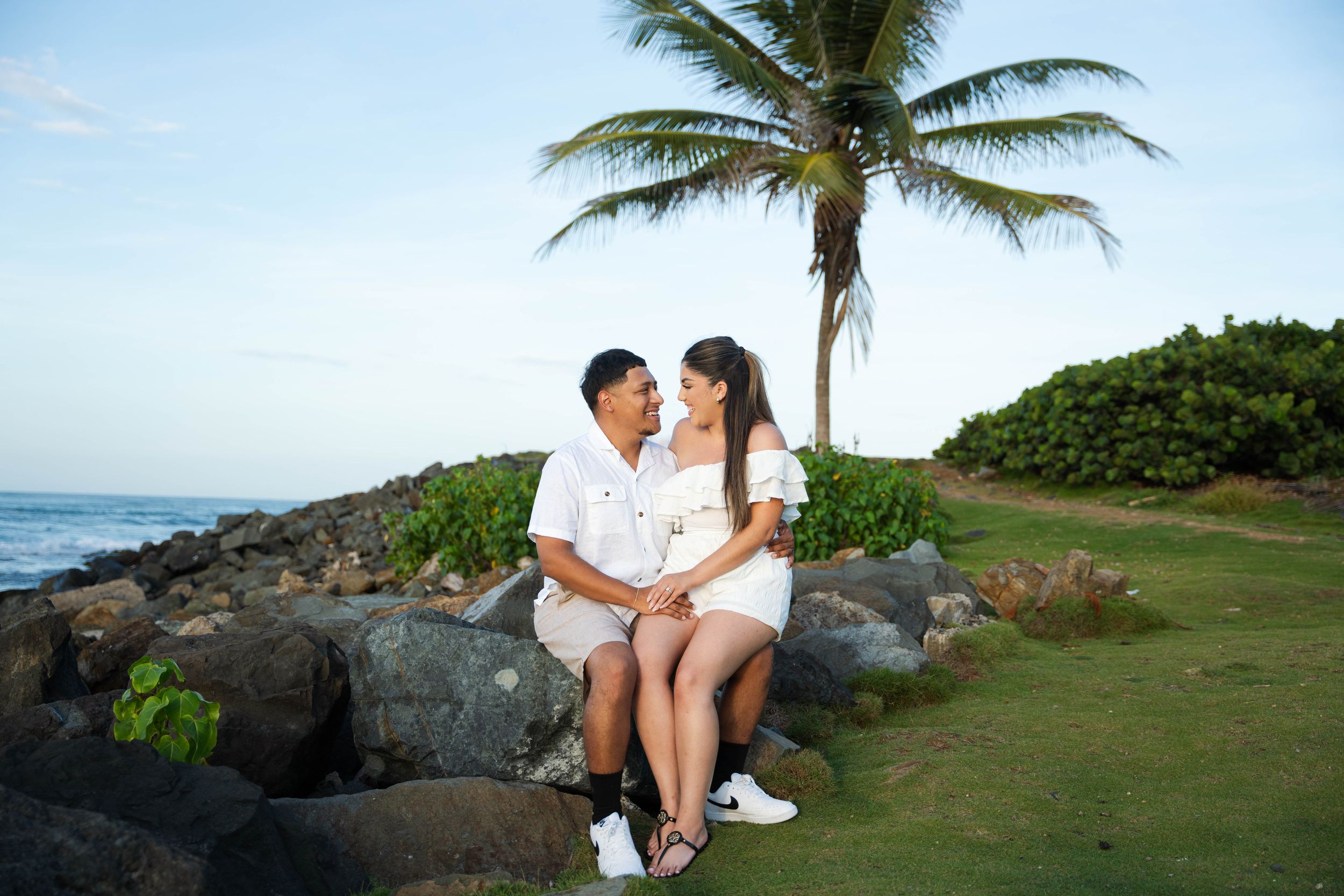 Couple sitting on rocky beach with palm tree during sunset photoshoot.