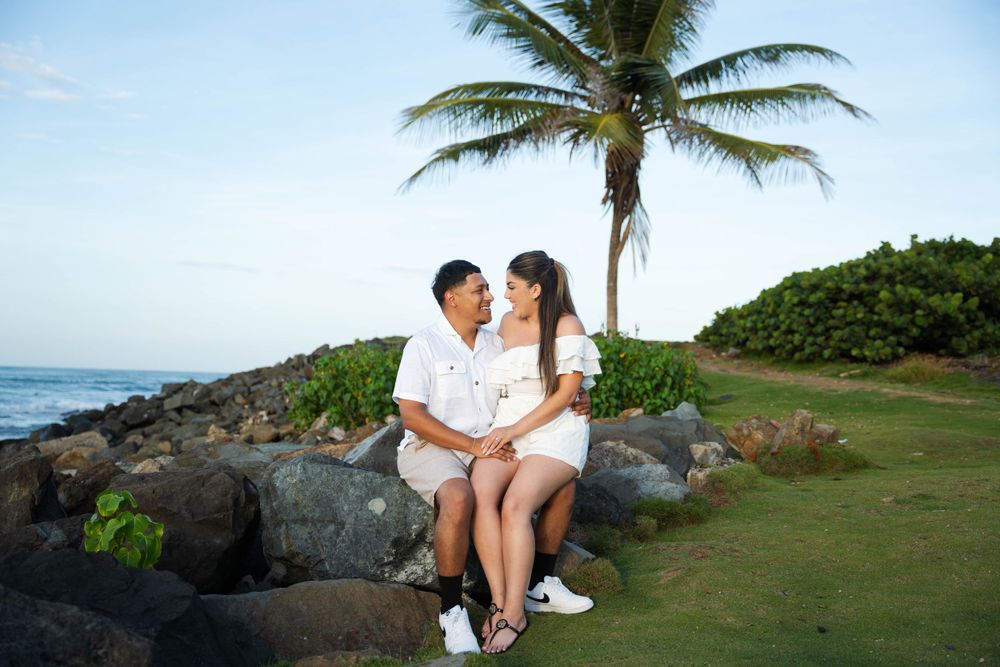 Couple sitting on rocky beach with palm tree during sunset photoshoot.
