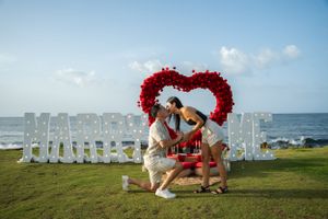 A romantic beach proposal setup with a large white letter sign and red heart-shaped arch by the ocean.