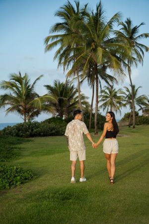 A couple walking hand in hand on a lush green lawn with tropical palm trees swaying in the background.