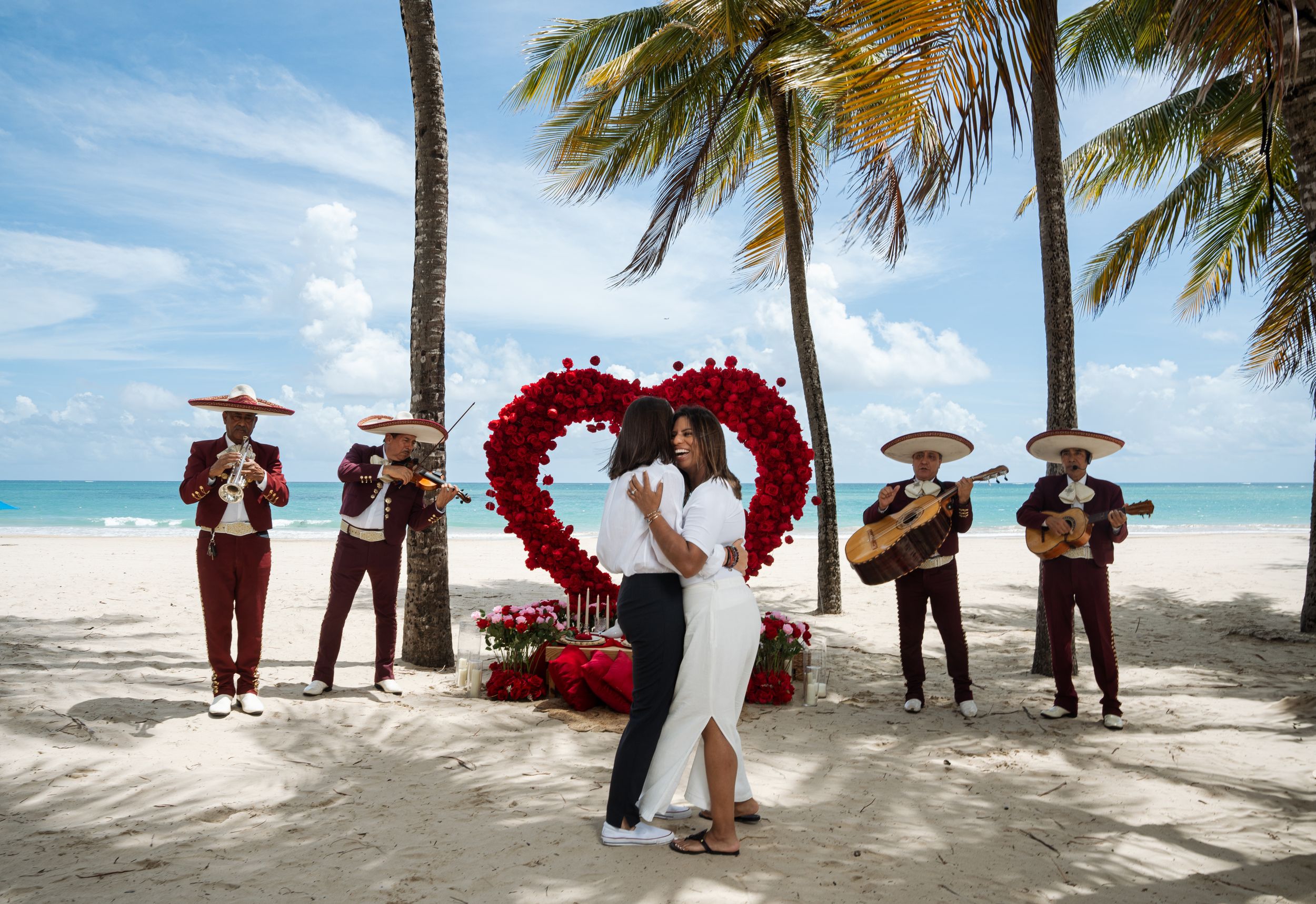 Beach wedding scene with mariachi band and heart-shaped arch made of red roses.