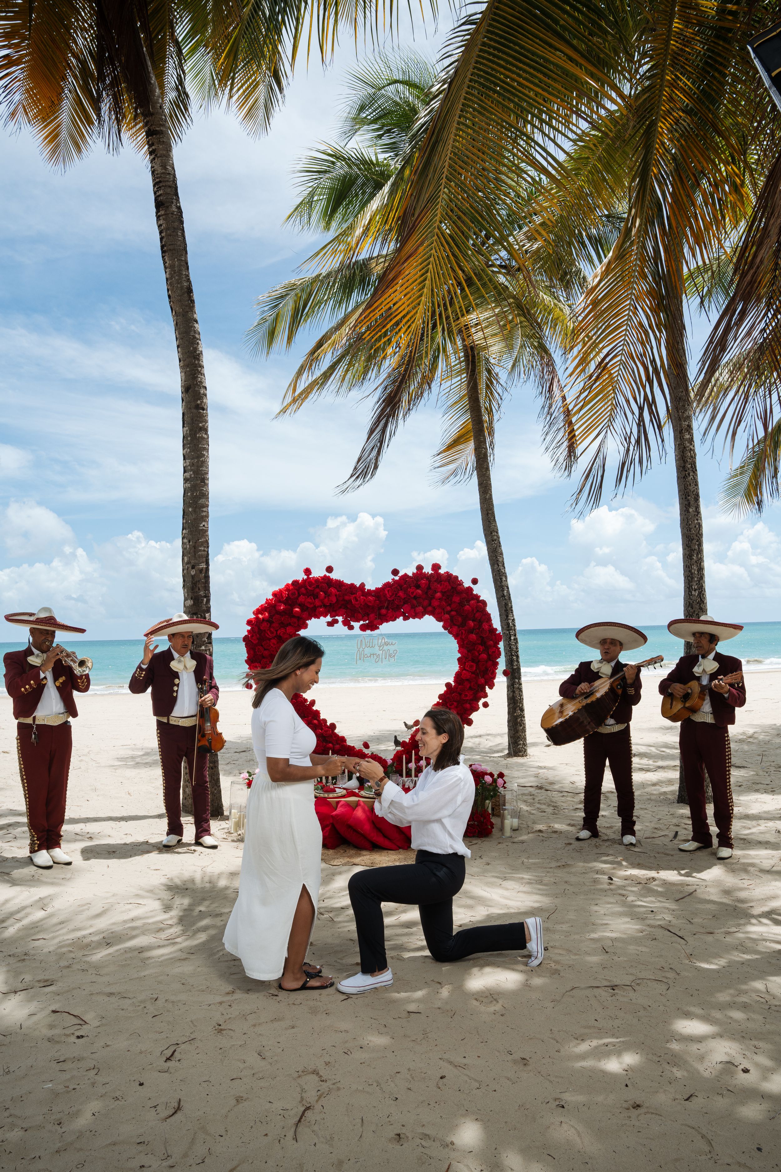 Proposal moment on beach with mariachi band and red heart-shaped flower arch.
