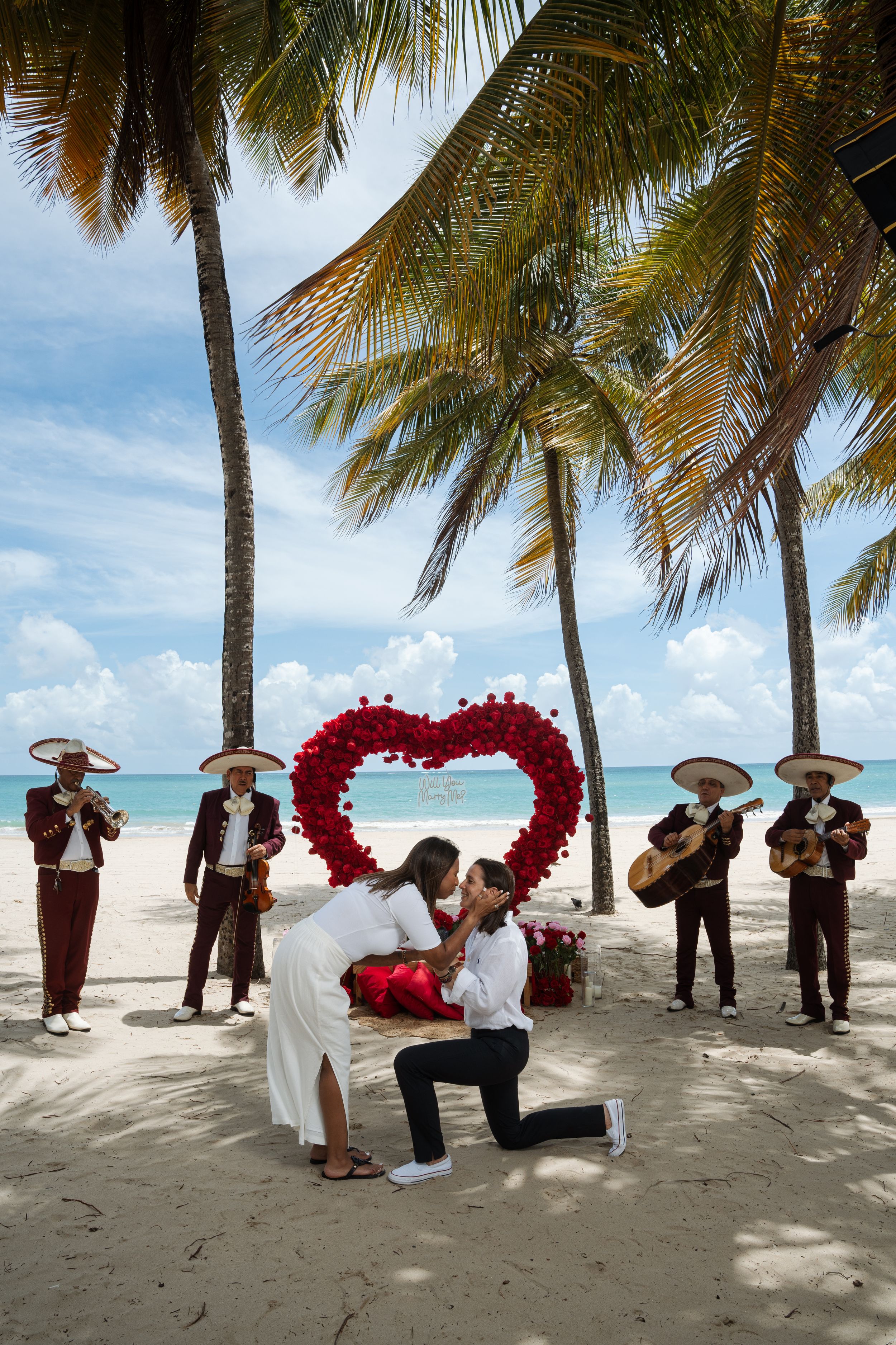 Beach proposal scene with traditional musicians and large red heart-shaped flower arrangement.