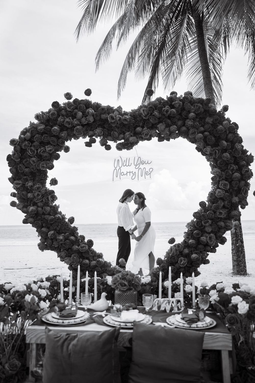 Black and white photo of romantic table setting under large balloon heart arch on beach.
