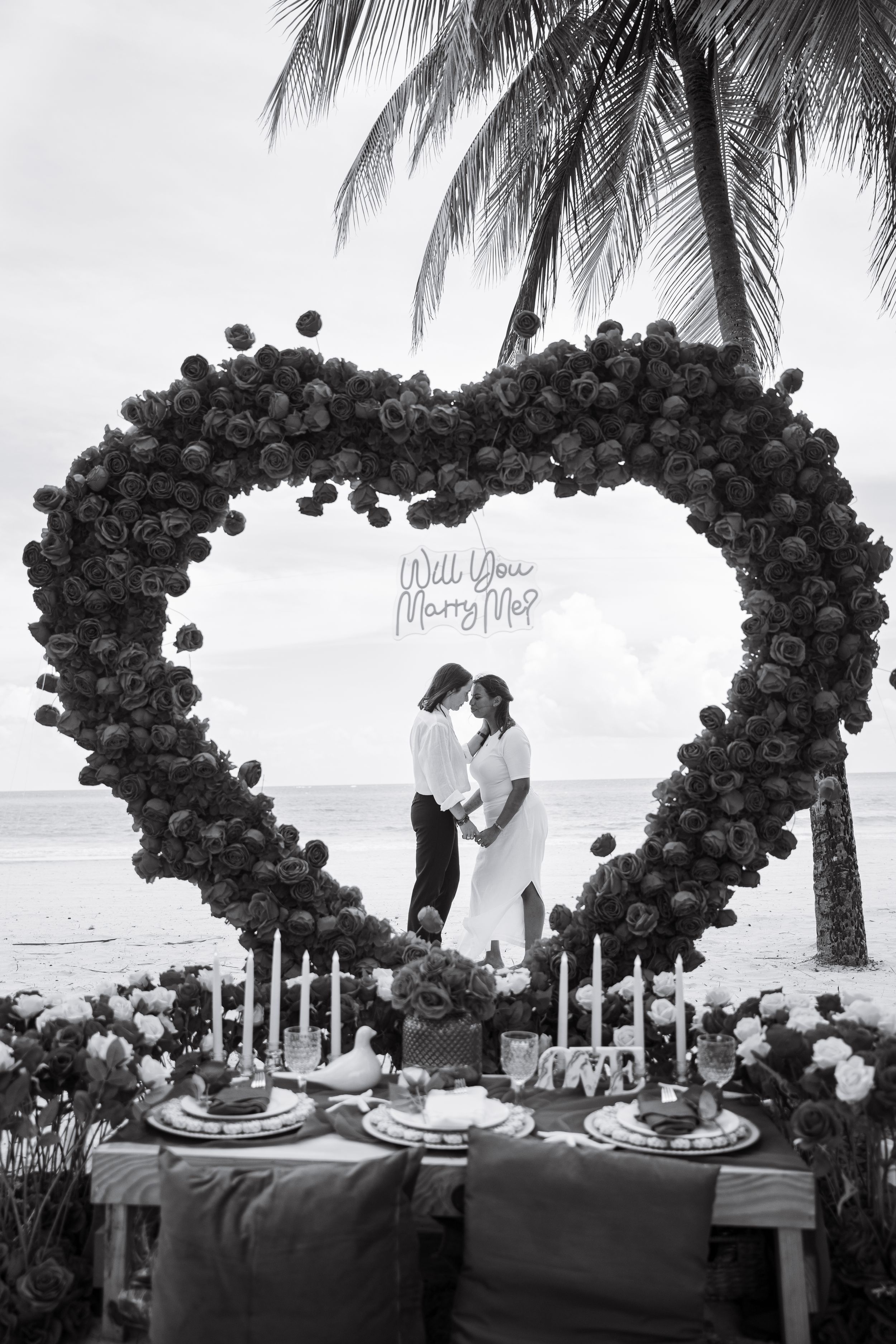 Black and white photo of romantic table setting under large balloon heart arch on beach.