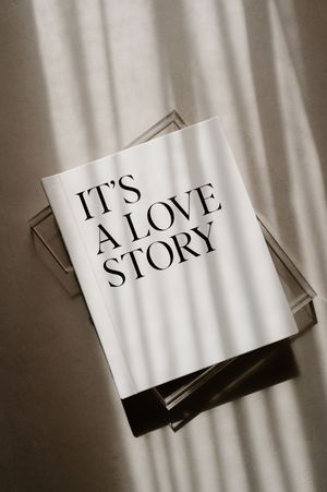 A white book or magazine cover with 'IT'S A LOVE STORY' text displayed against dramatic window light shadows.