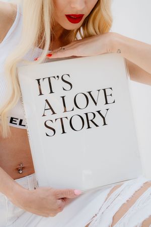 Someone holding a white book or magazine titled 'IT'S A LOVE STORY' while wearing white clothing.