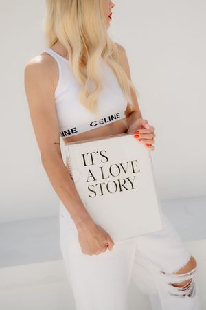 Woman in white athletic wear holds a white tote bag with 'It's A Love Story' text in elegant typography.