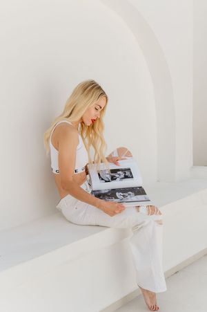 Someone in all white clothing sits in a minimalist white archway while looking through a photo album.