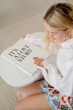 Reading a 'Love Story' book while wearing a white button-down shirt and patterned shorts in a cozy indoor setting.