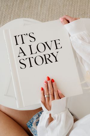 A white book with "IT'S A LOVE STORY" text held by hands with red nail polish and gold rings against a beige background.