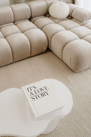 A white coffee table book titled 'It's A Love Story' sits near a plush beige tufted sectional sofa on neutral carpet.