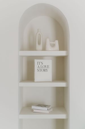 Minimalist white arched shelf with three levels displaying books and bottles, including one titled 'It's A Love Story'.