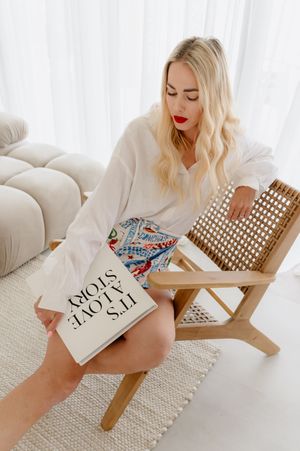 Woman in white sweater relaxes in modern living room with woven chair and It's About Slow book.