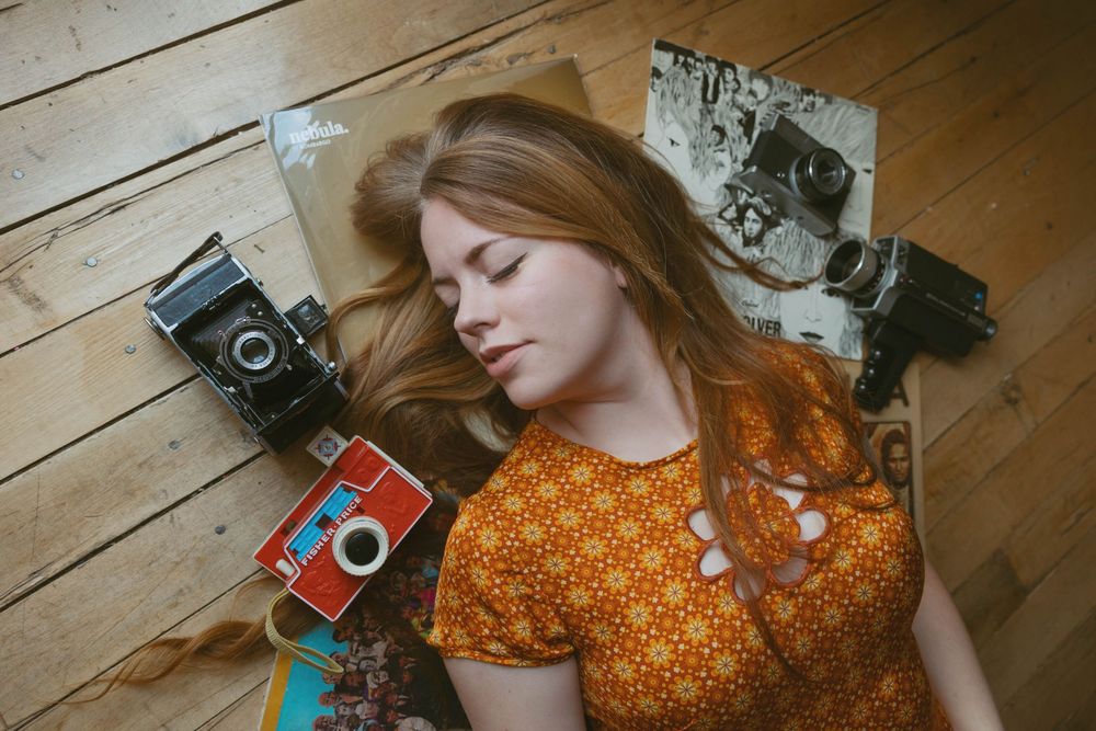 Young woman with red hair lying on wooden floor surrounded by vintage cameras in an orange floral dress.