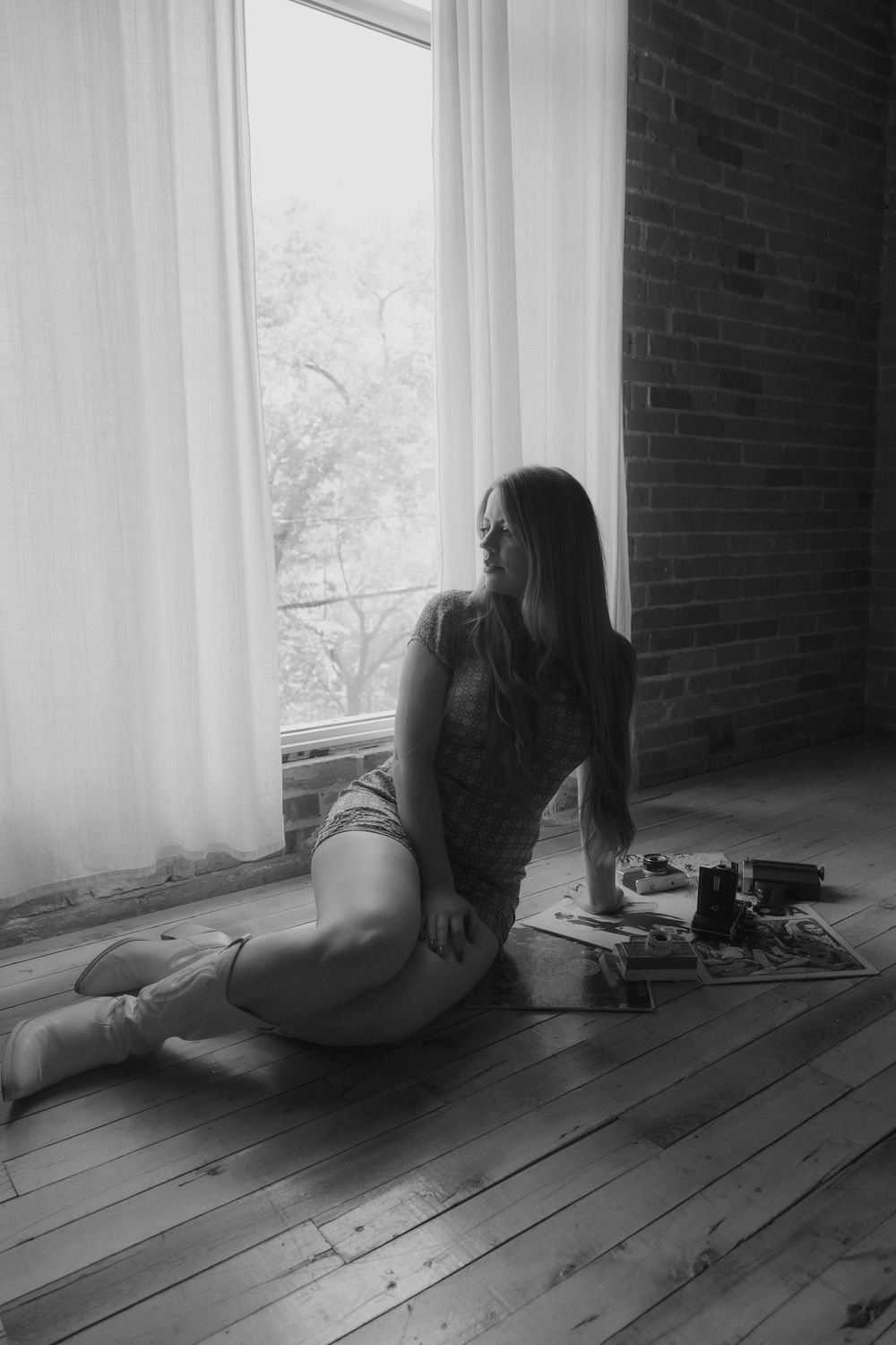 Black and white photo of a woman sitting on hardwood floor by a large window with sheer curtains and brick wall.