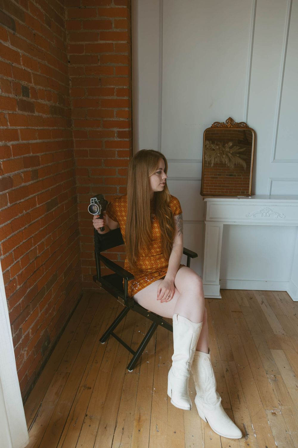 Young woman in orange dress and white cowboy boots sits on director's chair holding vintage camera near brick wall.