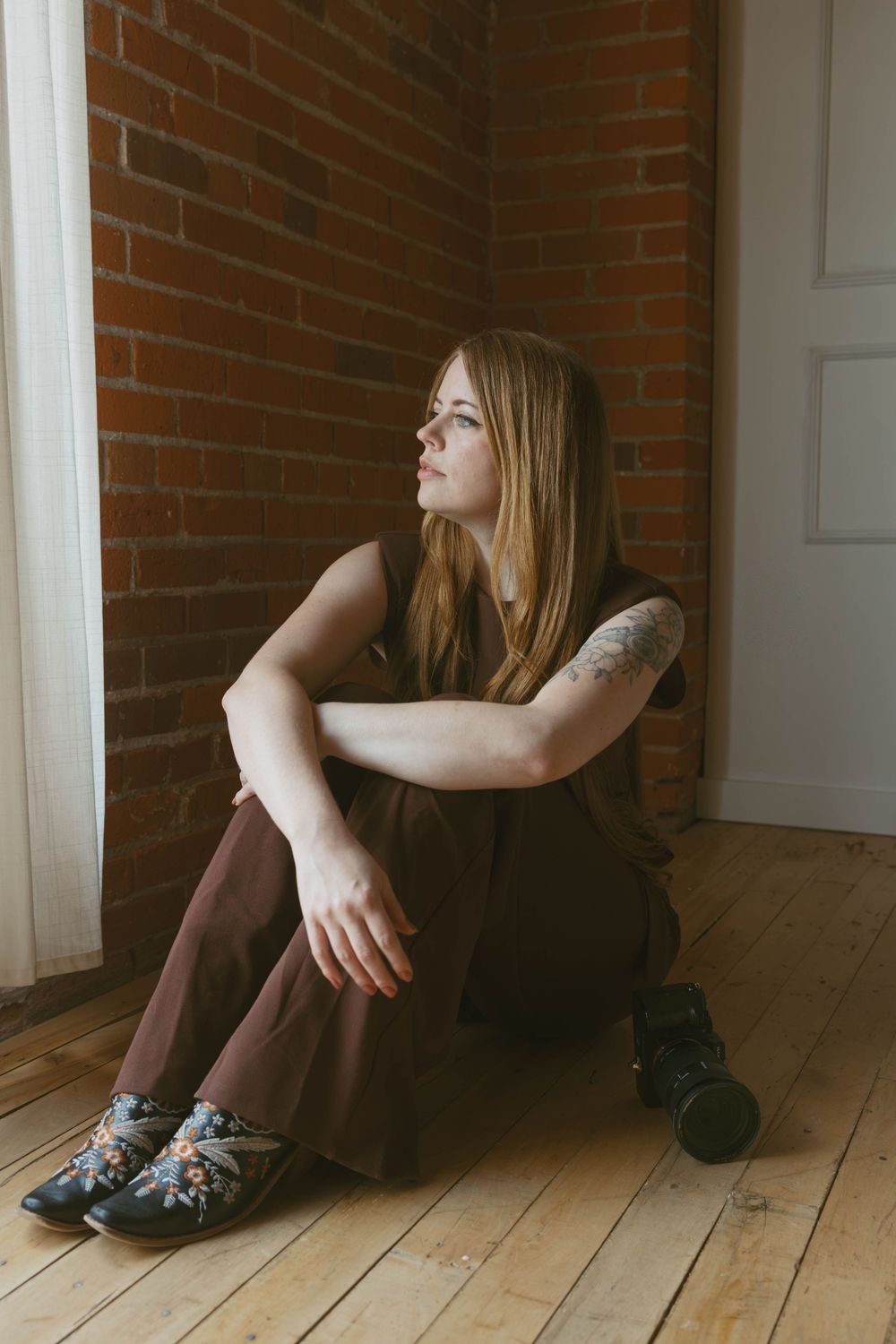 Young woman with long blonde hair and tattoo sits on wooden floor against brick wall, wearing brown outfit and floral shoes.