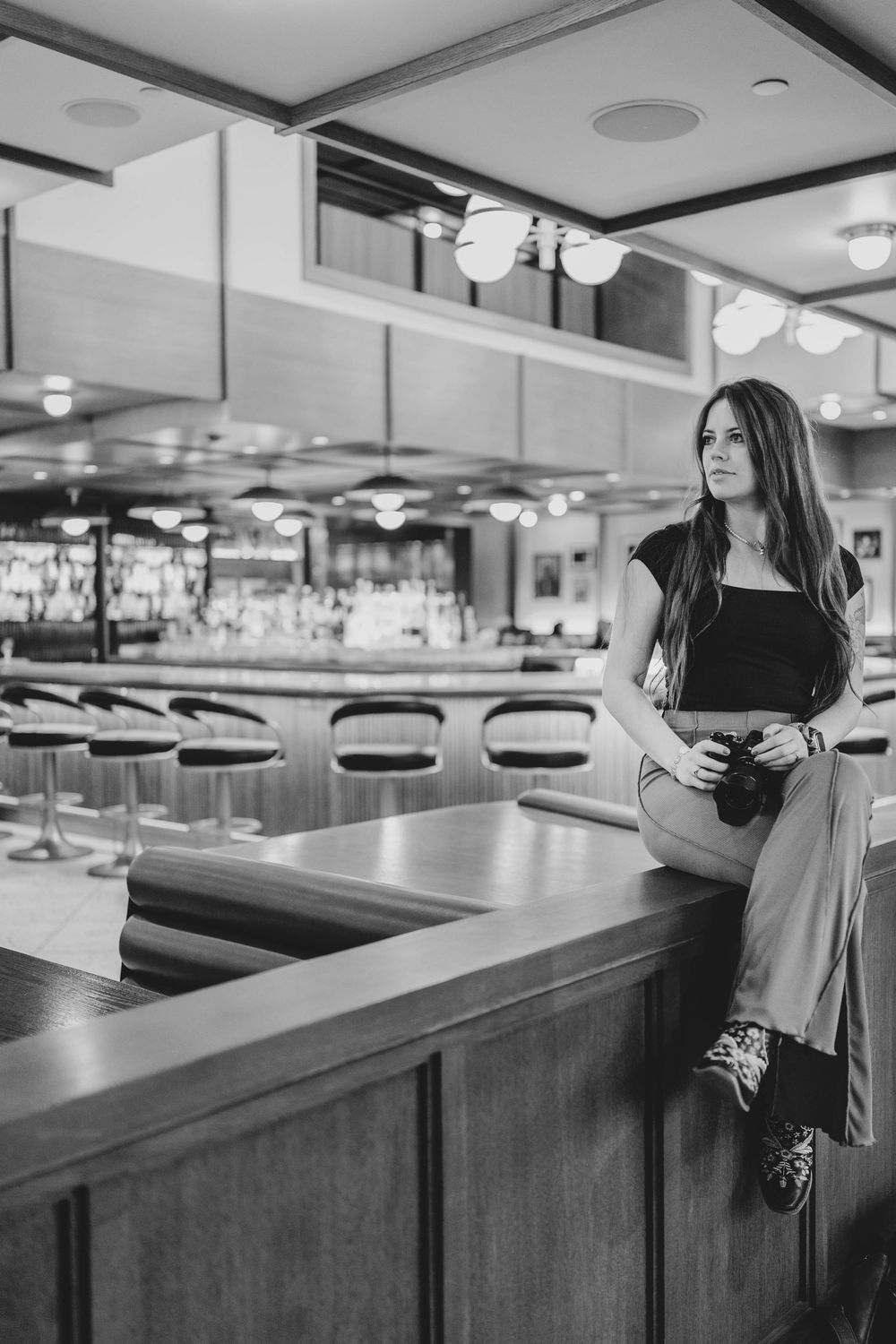 Woman in black top sits on wooden bar counter in elegant lounge, holding camera, black and white photo.
