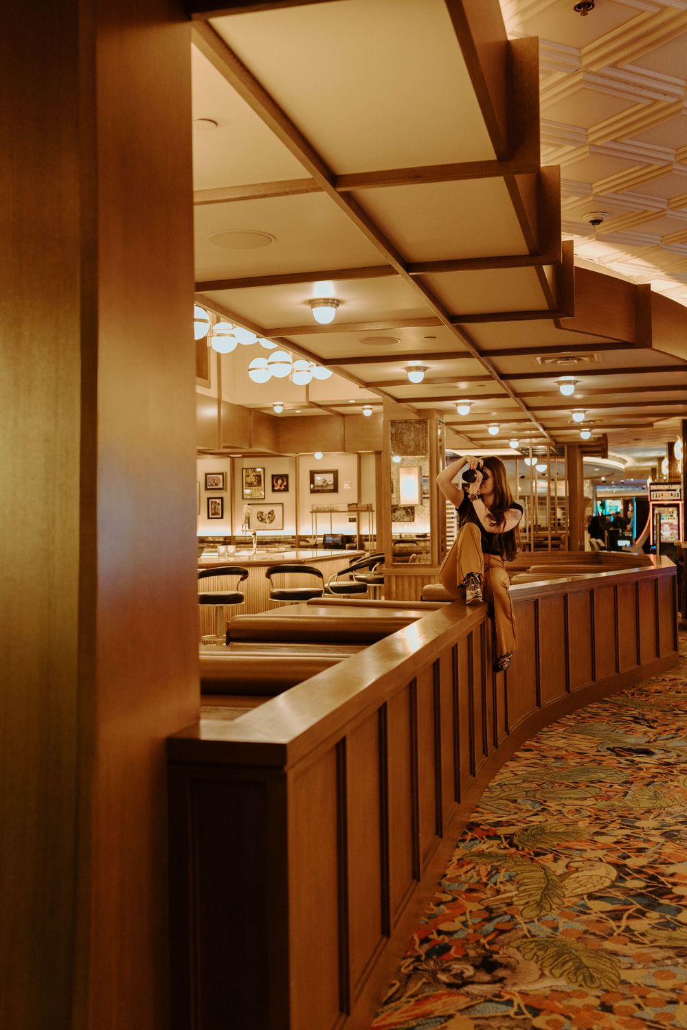 A woman stands in a warmly lit restaurant with wooden booths, ornate ceiling, and vibrant floral carpet.