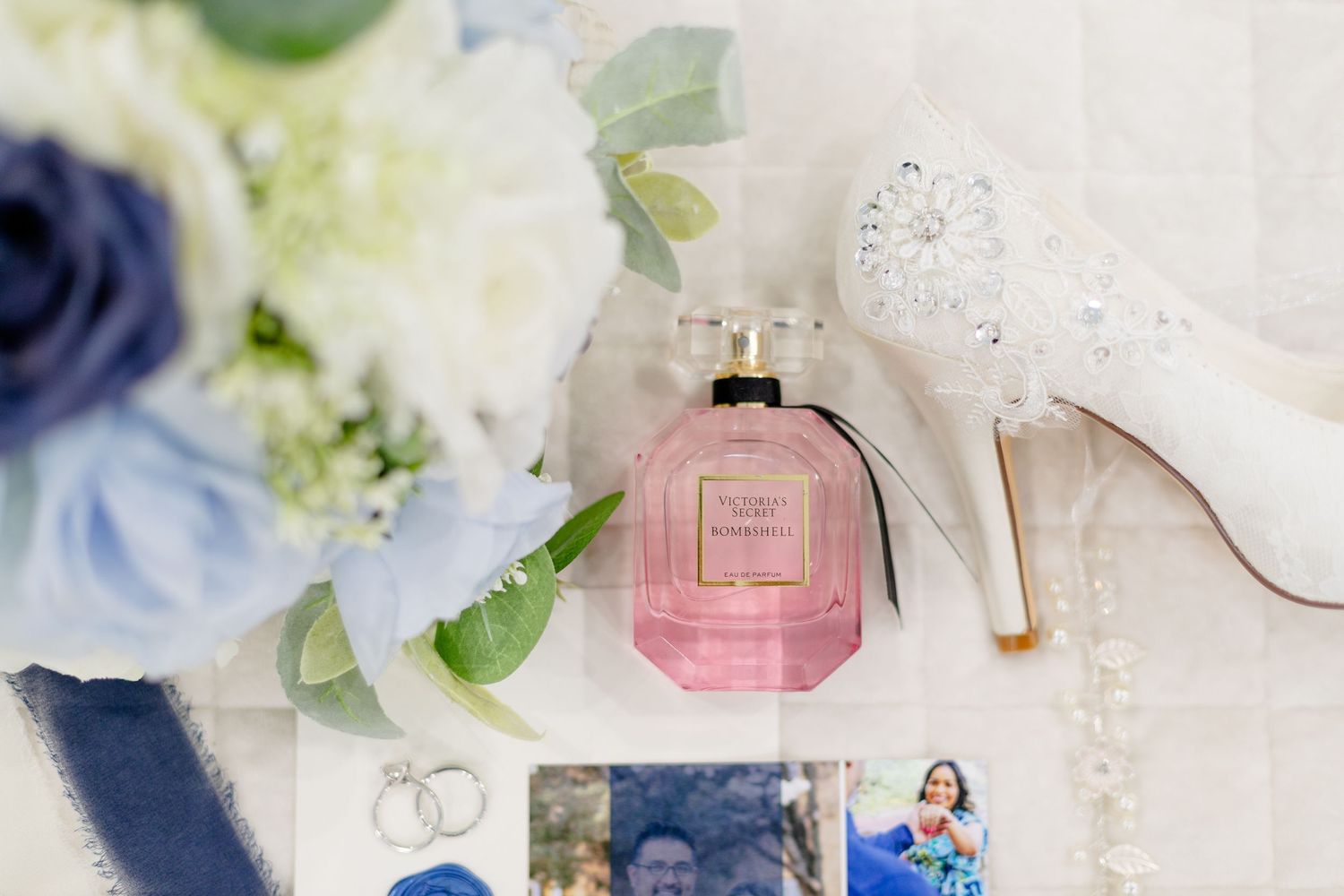 Wedding details featuring a pink perfume bottle, white heels, and wedding rings alongside blue flowers.