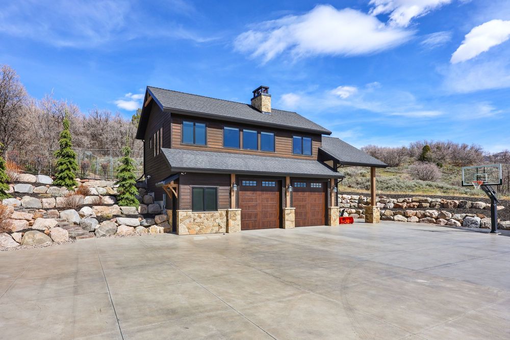 Rustic barn-style garage with dark wood siding, stone accents, dual garage doors, and mountain backdrop. Captured by Slate Media (best in UT