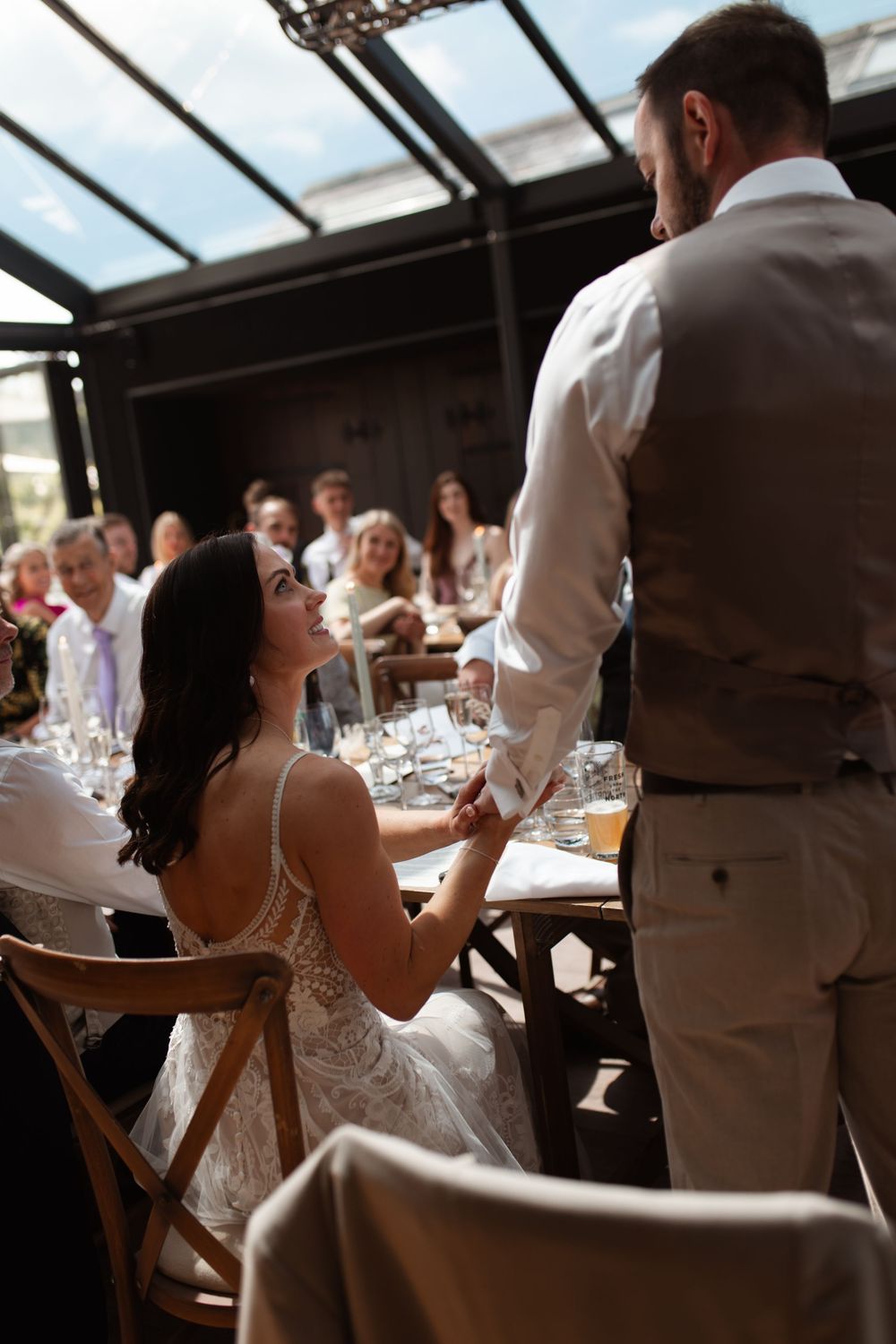 Bride in lace dress shares moment with server during wedding reception under glass ceiling conservatory.