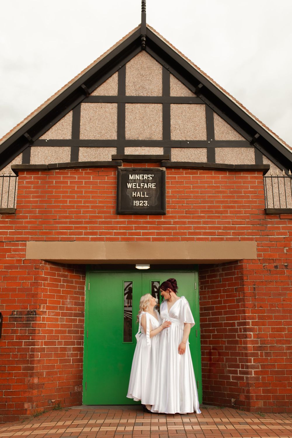 A couple in white dresses poses in front of a green door at Miners Welfare Hall, a brick Tudor-style building.