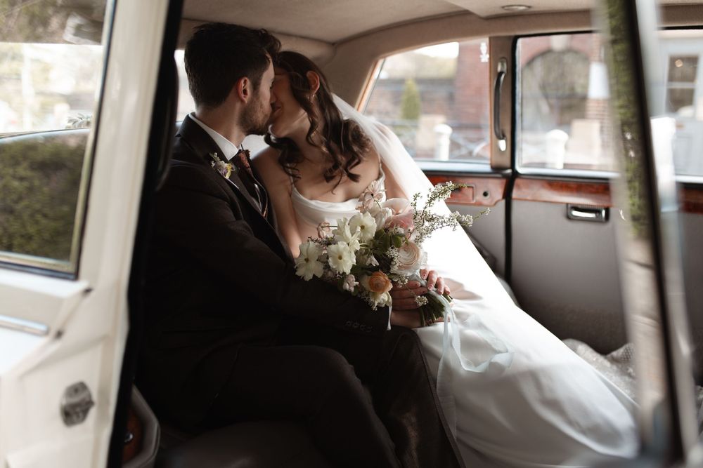 A tender romantic moment shared between newlyweds in the backseat of a classic car with a white floral bouquet.