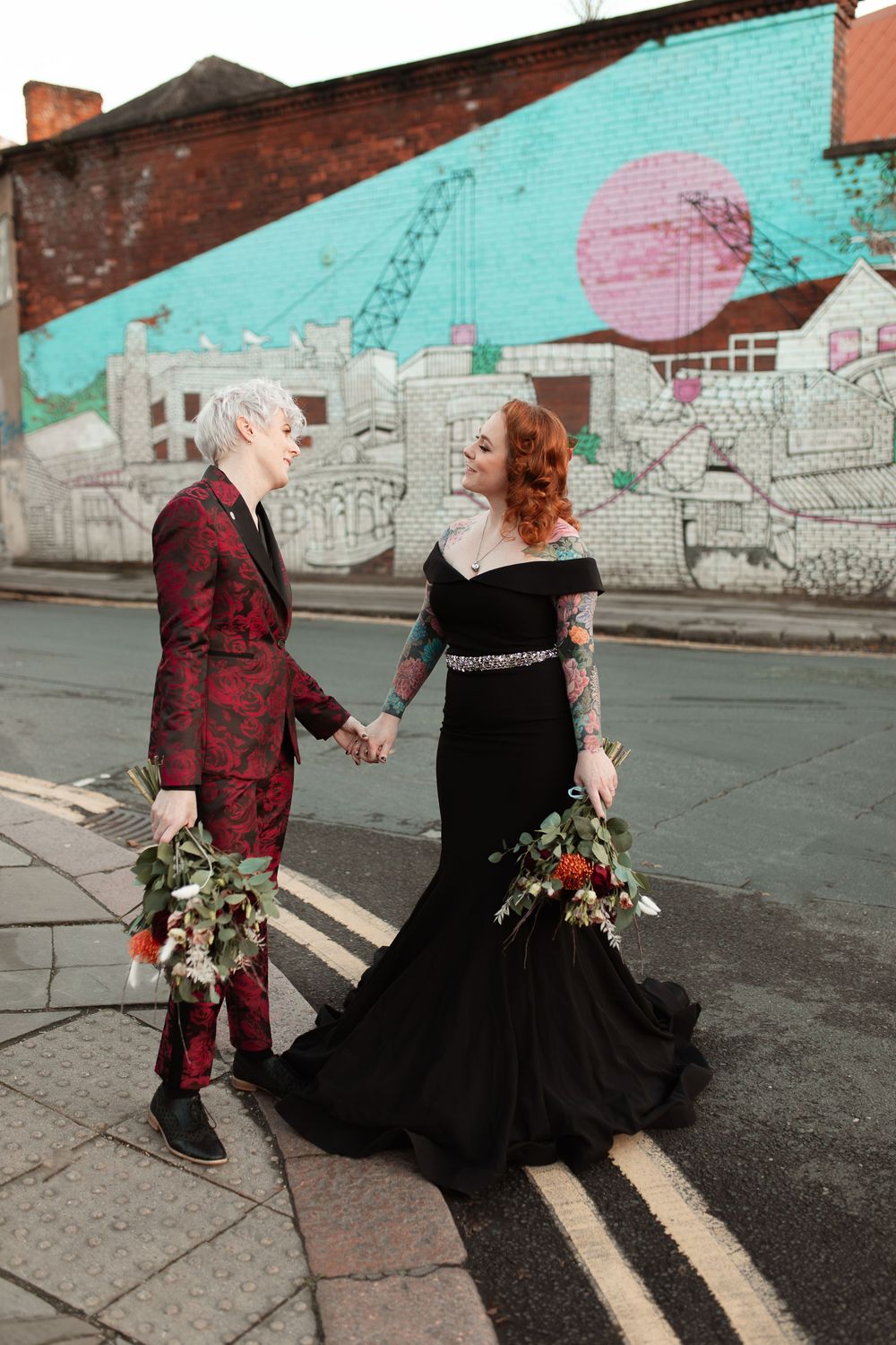 A couple in contrasting red and black outfits dances together on an urban street with a colorful mural wall behind them.