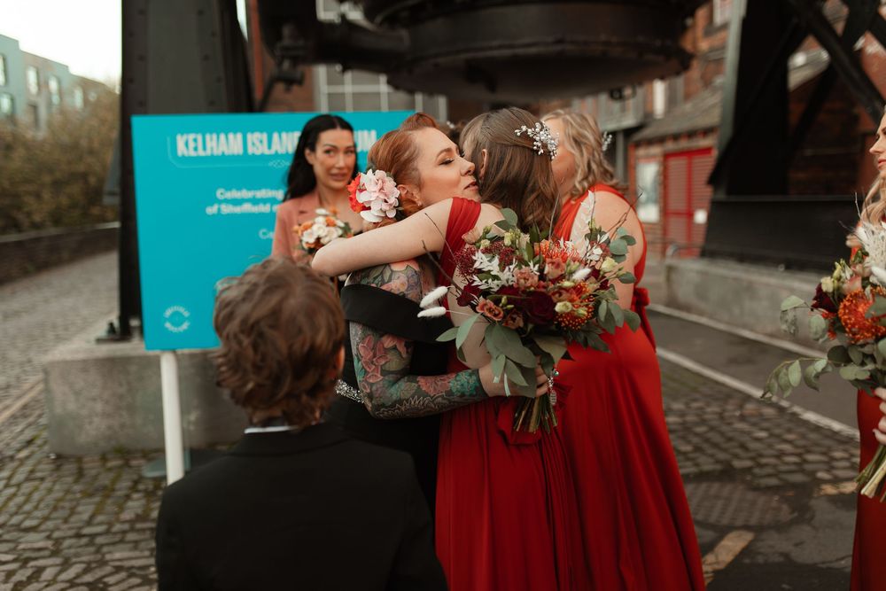 A group of people in formal attire gather around a sign, celebrating with bouquets and embraces on a cobblestone street.
