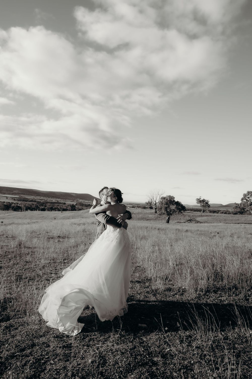Black and white photo of bride and groom embracing in a open field with rolling hills and cloudy sky. Kristielle Gibbons Photography.