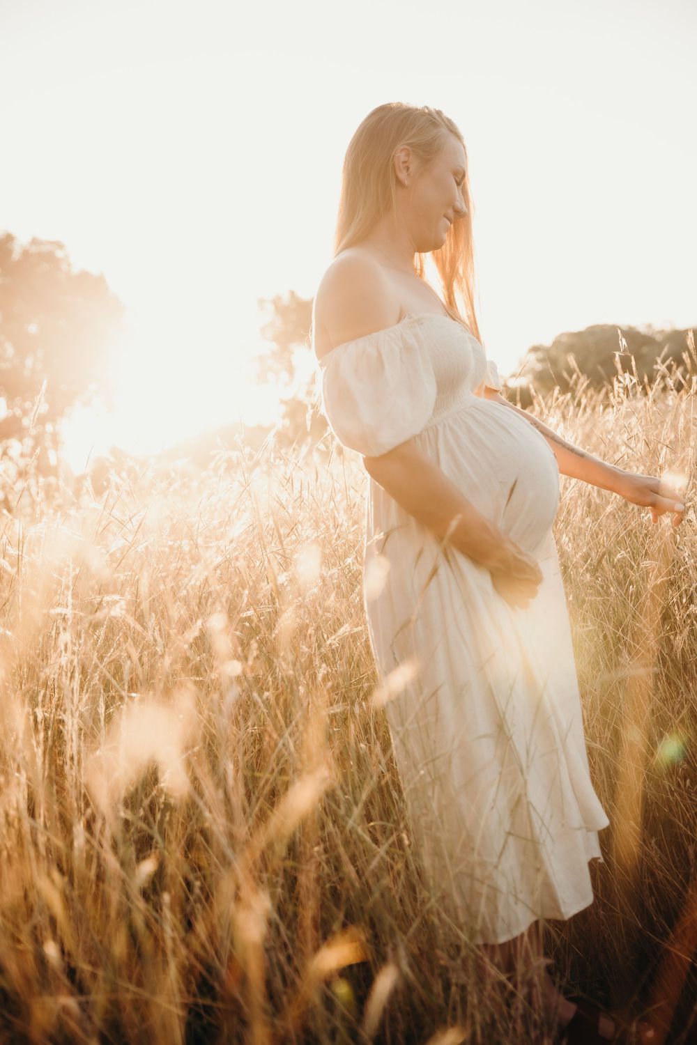 Pregnant woman in a white off-shoulder dress standing in a golden field during a maternity session.