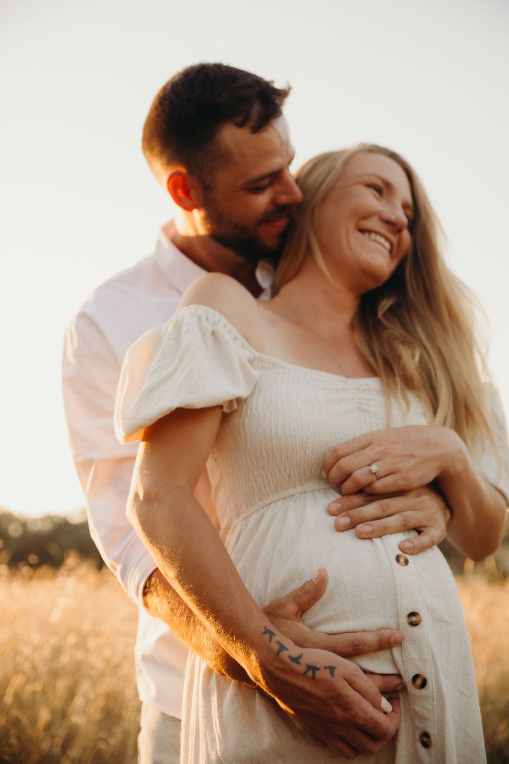 Couple embracing in a golden field at sunset, hands on pregnant belly during maternity photo session.