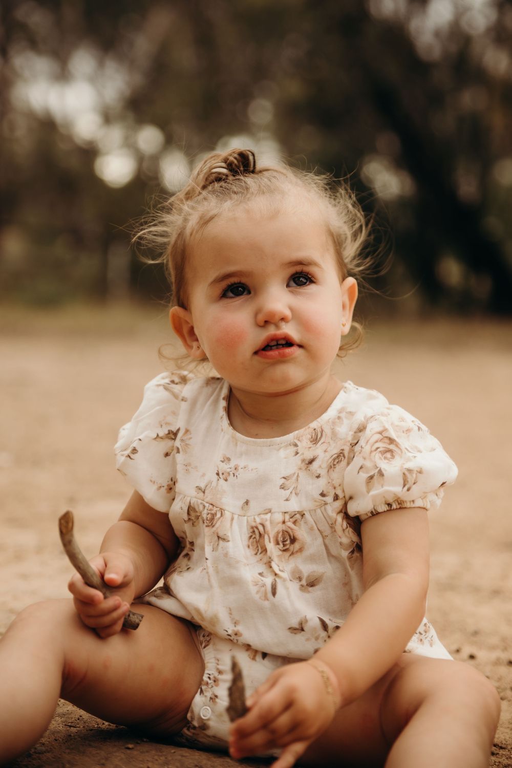 Cute toddler girl in white floral romper sitting on ground holding a stick outdoors.