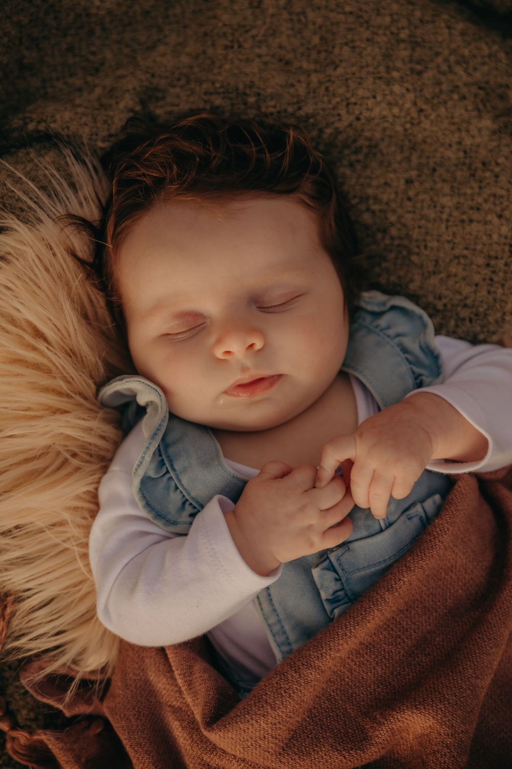 Sleeping newborn baby in denim vest wrapped in brown knit blanket on fur.