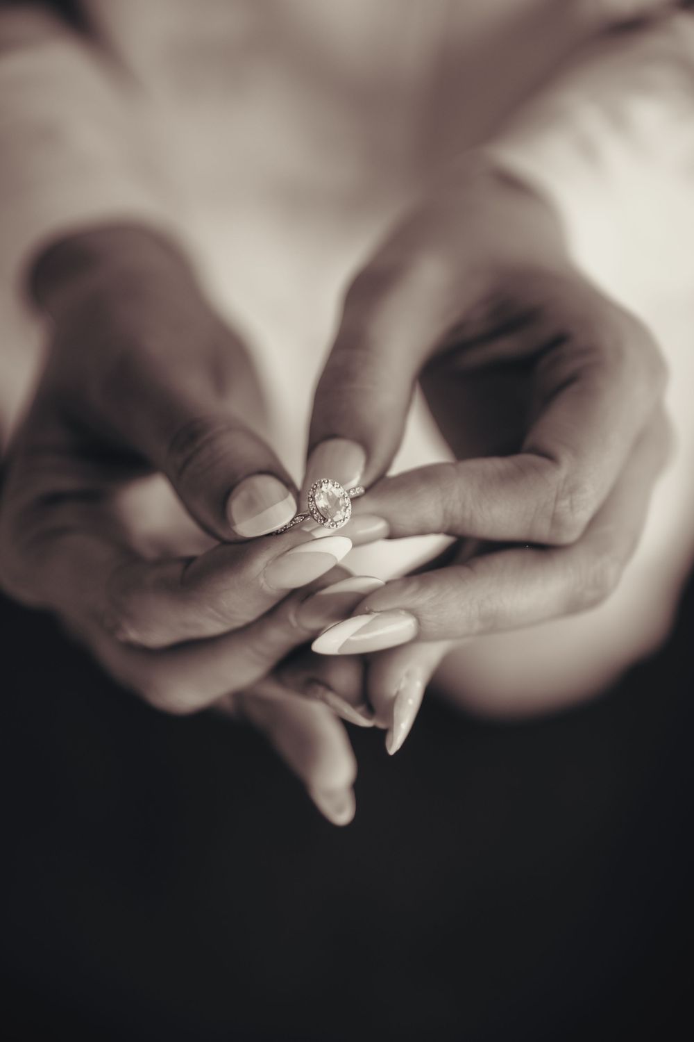 Sepia-toned close-up of a woman's hands with manicured nails holding a sparkling diamond engagement ring. Kristielle Gibbons Photography.