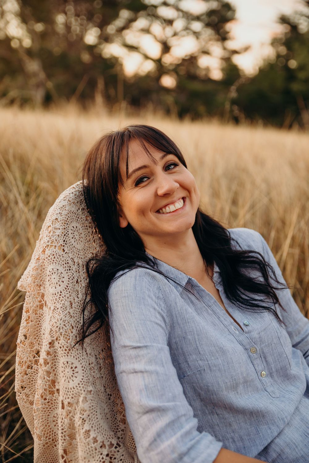 A dark-haired woman smiles warmly while relaxing in a golden grass field during a sunset portrait session.