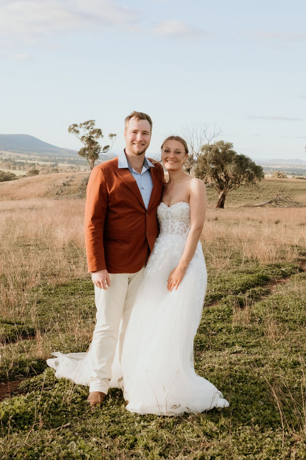 A wedding couple in a golden field, groom in rust jacket, bride in white lace gown, Australian landscape backdrop. Kristielle Gibbons Photo.