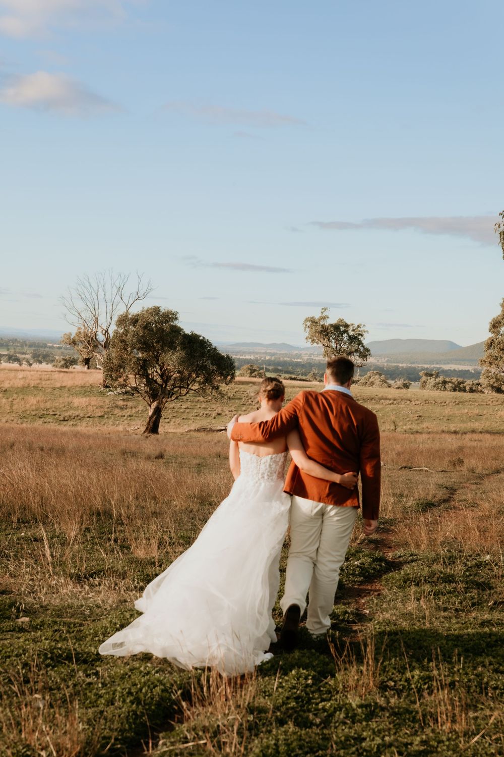 A bride and groom walk hand in hand through a golden rural landscape, viewed from behind, on their wedding day.
Kristielle Gibbons.